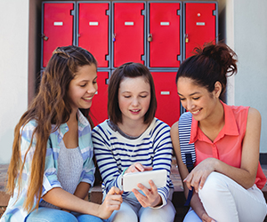 Three female students look at a smartphone in front of outdoor lockers
